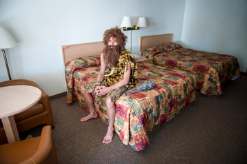 Lonely caveman traveler sitting indoors on the edge of double bed in a dingy motel room
