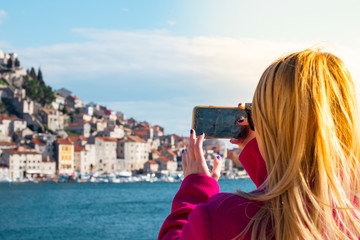 Fototapeta na wymiar Back view of a blonde woman in a pink coat holding a smartphone taking a photo, town of Sibenik, Croatia in the distance. Blue sea and stone houses
