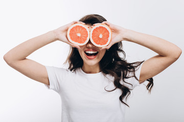 Smiling brunette girl in white t-shirt playing fool and covering her eyes with halves of grapefruit isolated white background