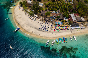 Fototapeta premium Top down aerial view of colorful boats and sunshades on a tropical beach on a small island fringed by a coral reef