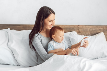 Motherhood. Mother and son at home sitting in bed taking selfie on smartphone smiling joyful