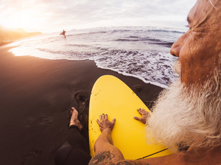 Happy surfer senior sitting on surfboard watching sunset time - Mature bearded man having fun on...