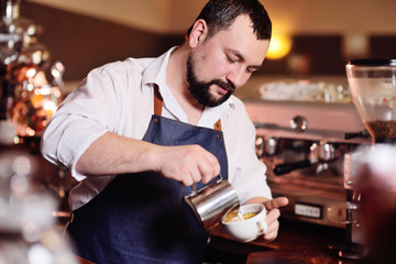 a handsome barista man in a white shirt and apron prepares fragrant coffee on the background of a...