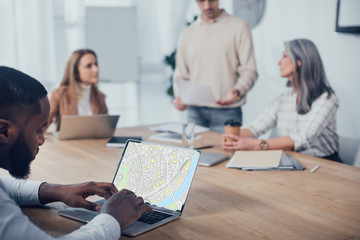 selective focus of african american man using laptop with map website and his colleagues talking on background