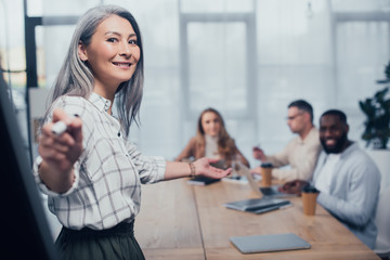 selective focus of smiling asian businesswoman pointing with marker and her multicultural colleagues on background
