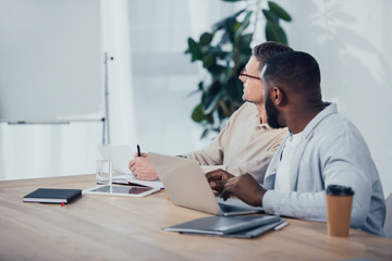 multicultural colleagues sitting at table and looking away in office