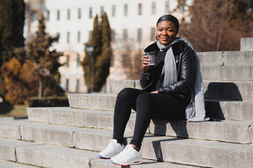 Portrait of a smiling young African American girl with pigtails with coffee walking in the street on a sunny day. Outdoor photo.