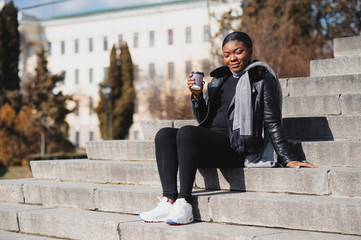 Portrait of a smiling young African American girl with pigtails with coffee walking in the street on a sunny day. Outdoor photo.