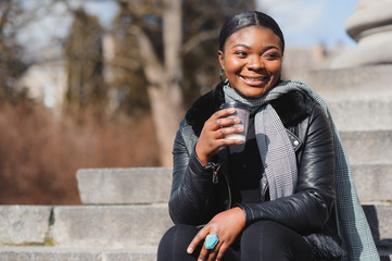 Portrait of a smiling young African American girl with pigtails with coffee walking in the street on a sunny day. Outdoor photo.