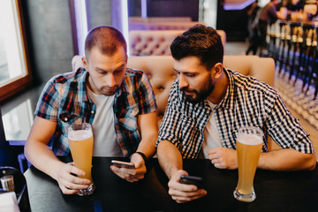 Just look at this photo. Two happy young men in casual wear drinking beer in pub while one of them holding smart phone and pointing it with smile