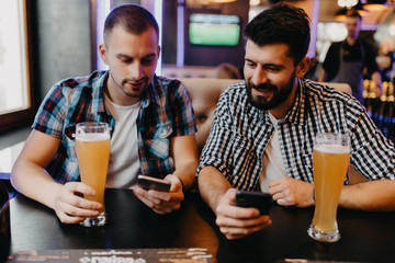 Just look at this photo. Two happy young men in casual wear drinking beer in pub while one of them holding smart phone and pointing it with smile
