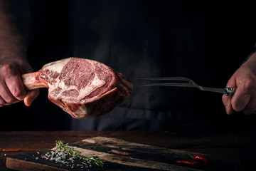 The chef's hands hold a raw Tomahawk a beef steak and a meat fork over a chopping Board