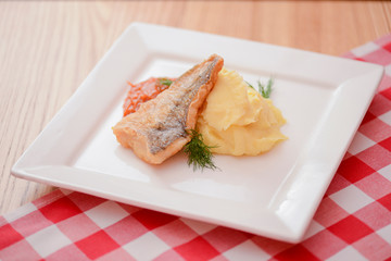 Fried fish filet with mashed potatoes and tomato sauce served on white square plate on wooden table.