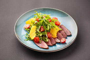Italian ham prosciutto salad with orange, cherry tomatoes and arugula served on a blue plate on dark background