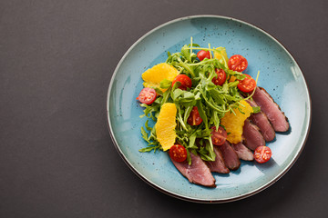 Italian ham prosciutto salad with orange, cherry tomatoes and arugula served on a blue plate on dark background