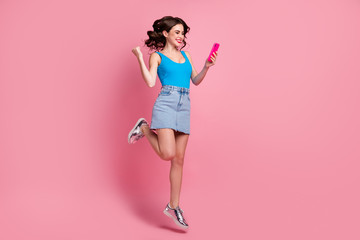 Full length profile photo of funny lady jump high hold telephone read good news raise fist astonished wear casual blue tank-top denim short skirt shiny shoes isolated pink color background