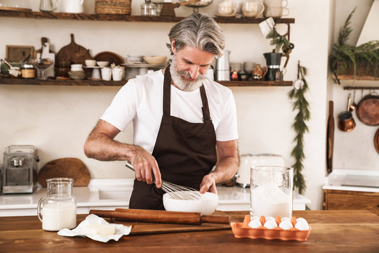 Image of concentrated gray-haired man in apron cooking dough for pastry