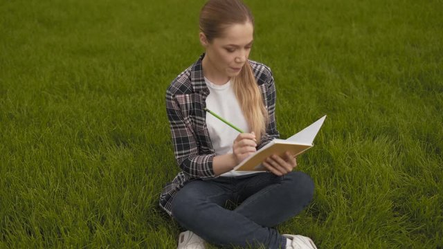teenager girl in a plaid shirt sits on the grass and takes notes in a notebook. 4K