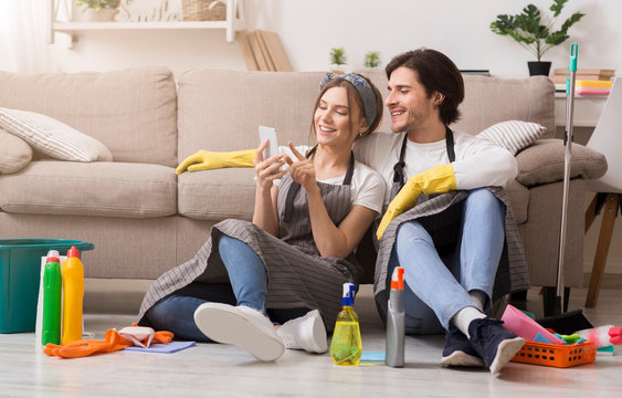 Smiling young couple relaxing with smartphone after cleaning apartment