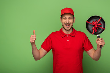 happy delivery man in red uniform holding frying pan with bow and showing thumb up on green