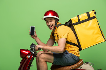 side view of happy delivery man in yellow uniform with backpack showing smartphone with blank screen on scooter isolated on green © LIGHTFIELD STUDIOS