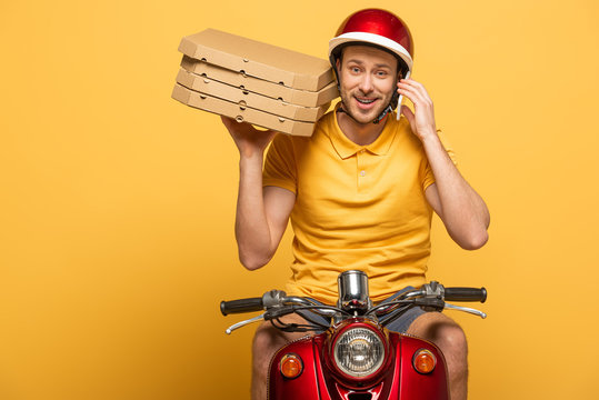 smiling delivery man in yellow uniform riding scooter with pizza boxes and talking on smartphone isolated on yellow