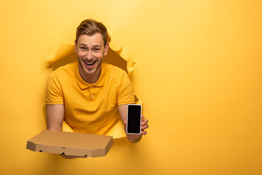 happy handsome man in yellow outfit holding smartphone and pizza box in yellow paper hole