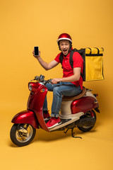 Fototapeta na wymiar happy delivery man with backpack on scooter holding smartphone on yellow background