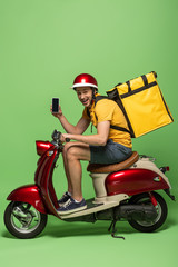 side view of happy delivery man in yellow uniform with backpack showing smartphone with blank screen on scooter on green © LIGHTFIELD STUDIOS