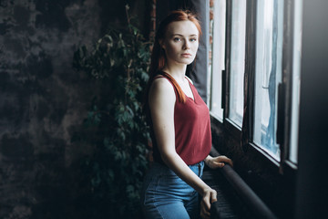 Close up portait of young sad, sensual, depressed, serious caucasian red headed gril with freckles standing near the window. Psychological condition concept