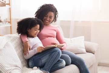 Beautiful pregnant afro woman and her adorable daughter reading book together