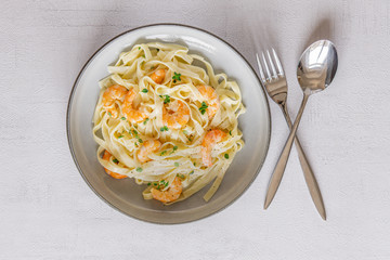 Italian pasta Tagliatelle in a creamy sauce with shrimp and persil on a plate, top view. Blue napkin. Fork and spoon inside. Copy space. Grey background. Concept: healthy food, tasty dinner. Top view.