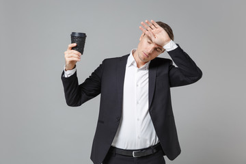 Naklejka premium Exhausted young business man in classic black suit shirt posing isolated on grey background. Achievement career wealth business concept. Mock up copy space. Hold cup of coffee or tea put hand on head.