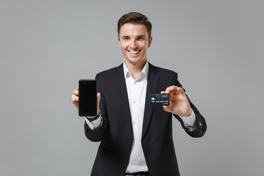 Smiling business man in classic suit shirt isolated on grey background. Achievement career wealth business concept. Mock up copy space. Hold mobile phone with blank empty screen, credit bank card.