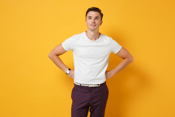 Fototapeta na wymiar Handsome young man guy 20s in white t-shirt posing isolated on yellow orange background in studio. People sincere emotions, lifestyle concept. Mock up copy space. Standing with arms akimbo on waist.