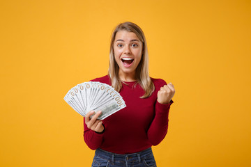 Excited young woman girl in casual clothes posing isolated on yellow orange background. People lifestyle concept. Mock up copy space. Hold fan of cash money in dollar banknotes doing winner gesture.