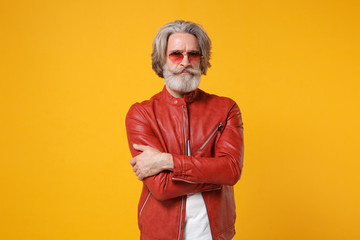 Serious elderly gray-haired mustache bearded man in red leather jacket, glasses posing isolated on yellow orange wall background. People lifestyle concept. Mock up copy space. Holding hands crossed.