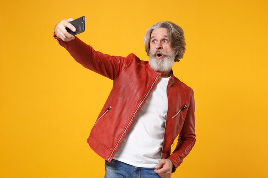 Shocked elderly gray-haired mustache bearded man in red leather jacket posing isolated on yellow orange background. People lifestyle concept. Mock up copy space. Doing selfie shot on mobile phone.