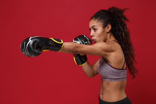 Side view of strong african american sports fitness boxer woman in sportswear working out isolated on red background. Sport exercise healthy lifestyle concept. Make boxing exercises in boxing gloves.