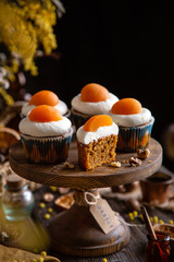 easter carrot cupcakes with white cream and slice of apricot on top (egg style decorated cupcakes) on wooden cake stand on rustic table with willow, oranges, nuts, spices