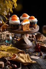 easter carrot cupcakes with white cream and slice of apricot on top (egg style decorated cupcakes) on wooden cake stand on rustic table with willow, oranges, nuts, spices