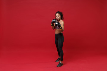Confident young african american sports fitness boxer woman in sportswear working out isolated on red wall background. Sport exercise healthy lifestyle concept. Make boxing exercises in boxing gloves.