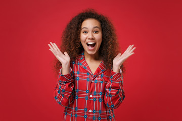 Surprised young african american girl in pajamas homewear posing resting at home isolated on red background. Relax good mood lifestyle concept. Mock up copy space. Keeping mouth open, spreading hands.