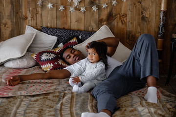 Caring loving young mixed race father carefully communicates with his beautiful little daughter lying on the bed in a cozy house. Young happy father rejoices and admires cute daughter lying on sofa