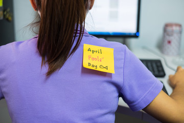 Back of young woman office with a yellow sticky note attached with the text Kick me. happy april...