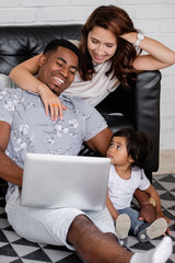 Beautiful young family african american dad asian mom and charming little daughter watching tv shows and cartoons using laptop smartphone and wi fi internet sitting on floor in modern interior