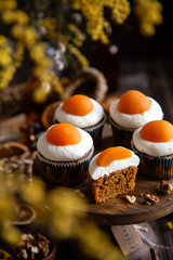 easter carrot cupcakes with white cream and slice of apricot on top (egg style decorated cupcakes) on wooden cake stand on rustic table with willow, oranges, nuts, spices