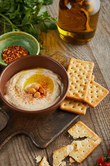Selective focus of crackers, parsley, bowls with paprika and hummus on cutting board on wooden background