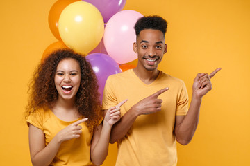 Laughing friends couple african american guy girl in casual clothes isolated on yellow background. Birthday holiday party concept. Celebrate hold colorful air balloons pointing index fingers up aside.