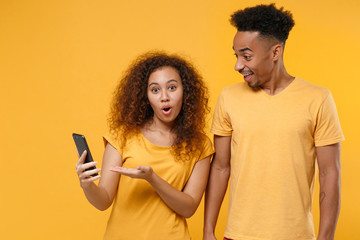 Shocked young friends couple african american guy girl in casual clothes isolated on yellow background. People sincere emotions lifestyle concept. Mock up copy space. Pointing hand on mobile phone.